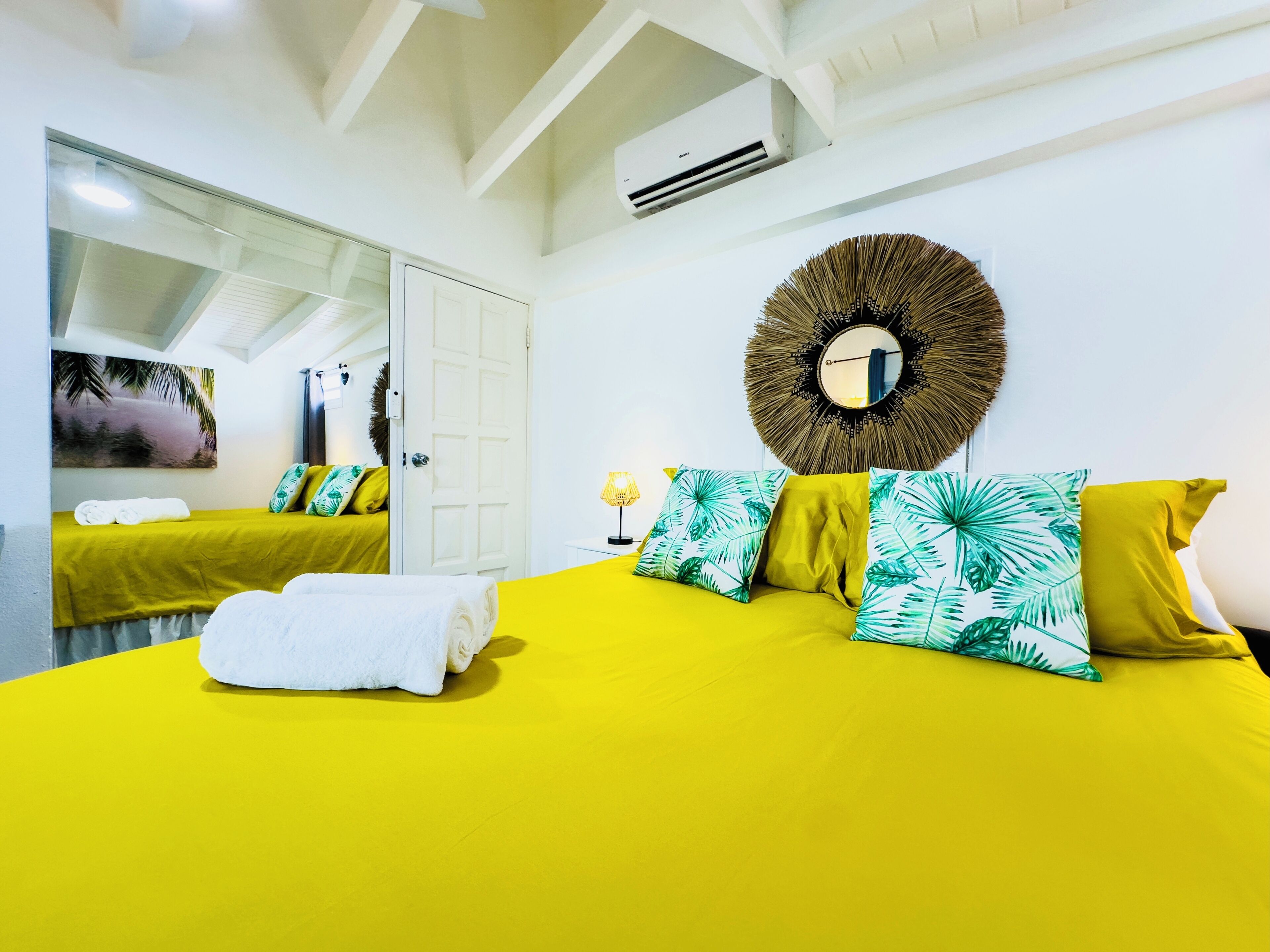 Una habitación luminosa de color amarillo con una cama, almohadas, un espejo y un equipo de aire acondicionado.