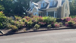 Uma casa de dois andares com fachada de pedra e várias janelas, rodeada por um jardim com diversas plantas floridas.