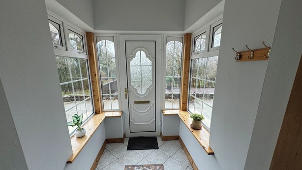 Um corredor com piso de azulejos, um banco de madeira, uma porta com painel de vidro decorativo e janelas com plantas.