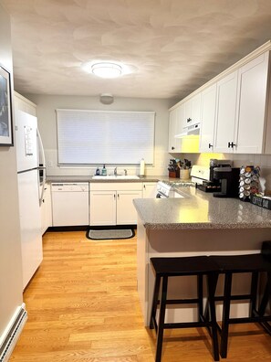 A kitchen with white cabinets, a granite countertop, a coffee bar, and a window with blinds.