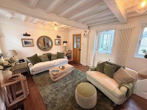 Ein Wohnzimmer mit einer Couch, einem Hocker und einem Couchtisch. Es befindet sich ein Spiegel, eine Lampe und ein Fenster mit Vorhängen dort.