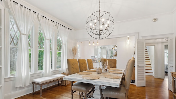 A dining room with a large wooden table, white curtains, and a chandelier.