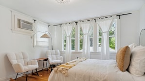 A bedroom with a large bed, two rocking chairs, a small table, and a window with curtains.