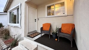 A house entrance with a white door, two wicker chairs with orange cushions, a potted plant, and a wall-mounted light fixture.