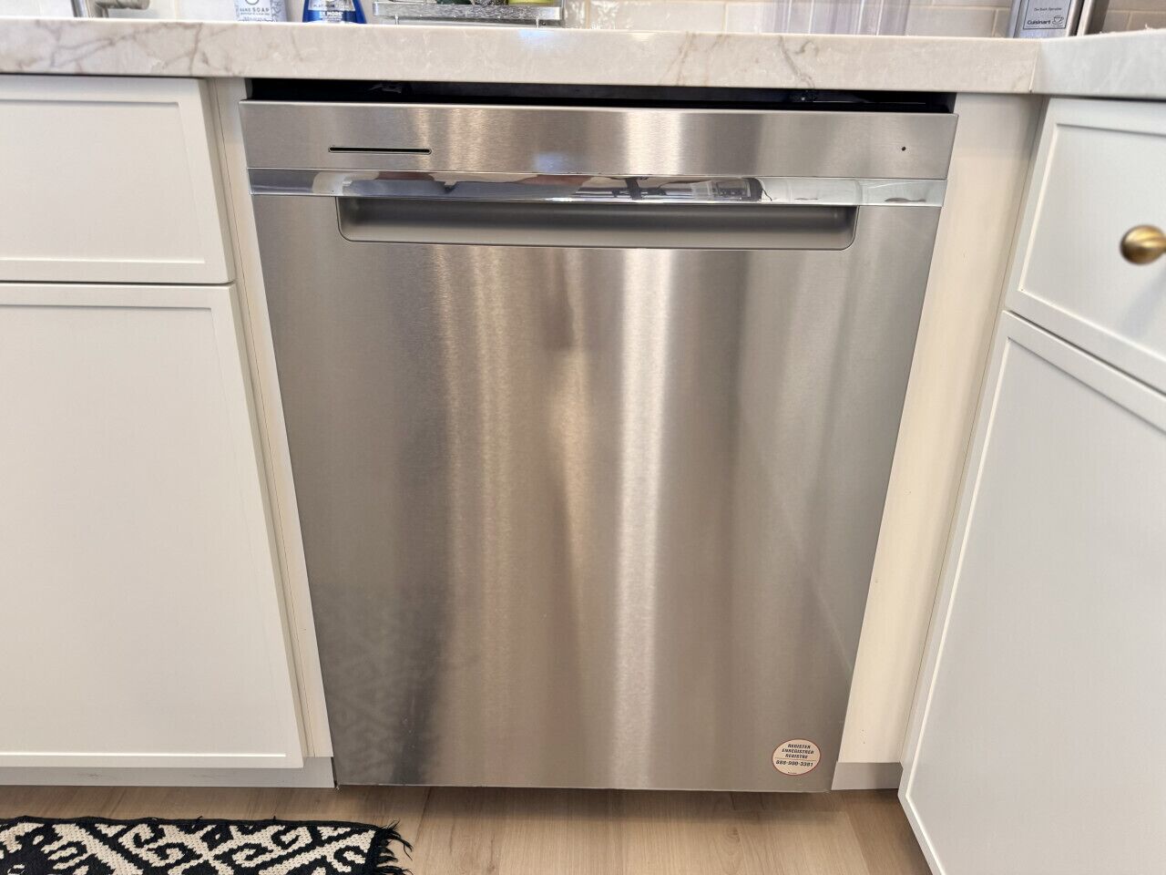 En diskmaskin i rostfritt stål i ett kök med vita skåp och en bänkskiva i marmor.