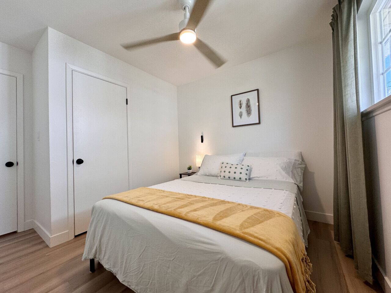 A bedroom with a bed, a ceiling fan, a nightstand, a framed picture on the wall, and a window with curtains.