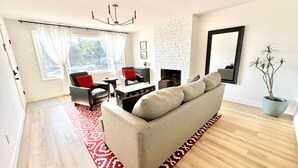 A spacious living room with a beige sofa, a red and white patterned rug, a fireplace, and a large window with curtains.