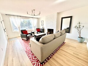 A spacious living room with a beige sofa, a red and white patterned rug, a fireplace, and a large window with curtains. - 4 Bed 2 Bath House Near Everything in San Diego.  (San Diego)