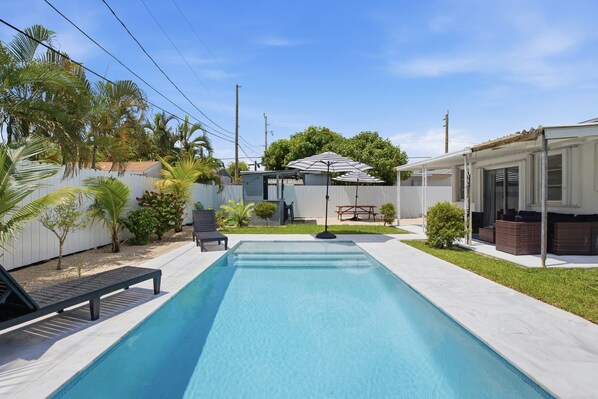 Una piscina rectangular con plataforma blanca, rodeada por una valla blanca, palmeras y un pequeño patio con mesa y sillas.