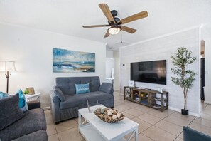 A living room with a grey sofa, a coffee table with decorative items, a ceiling fan, a painting on the wall, and a flat-screen TV.
