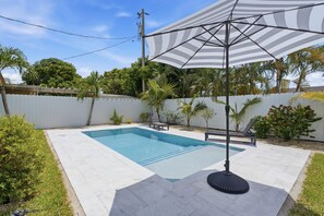 A swimming pool with a tiled deck, surrounded by a white fence, palm trees, and a white umbrella.