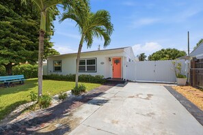 Una casa de un piso con puerta roja, cerca blanca en el jardín y una palmera.