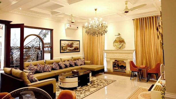 A spacious living room with a large sectional sofa, a chandelier, a fireplace, and a view of an outdoor area through a window. - Spanish Villa/5 BHK/Raya Golf course & Cinema 2Kms (Lahore)