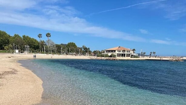 A beach with clear shallow waters, a sandy shore, and a building in the background. - Large Studio 100m from the beach (Toulon)