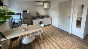 Et kompakt køkken med mikroovn, køleskab og vaskemaskine. Spisebord med stole og en lille plante.