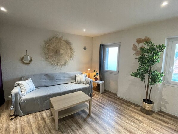 Een woonkamer met een grijze bank, een houten salontafel, een klein bijzettafeltje met een lamp en een potplant.