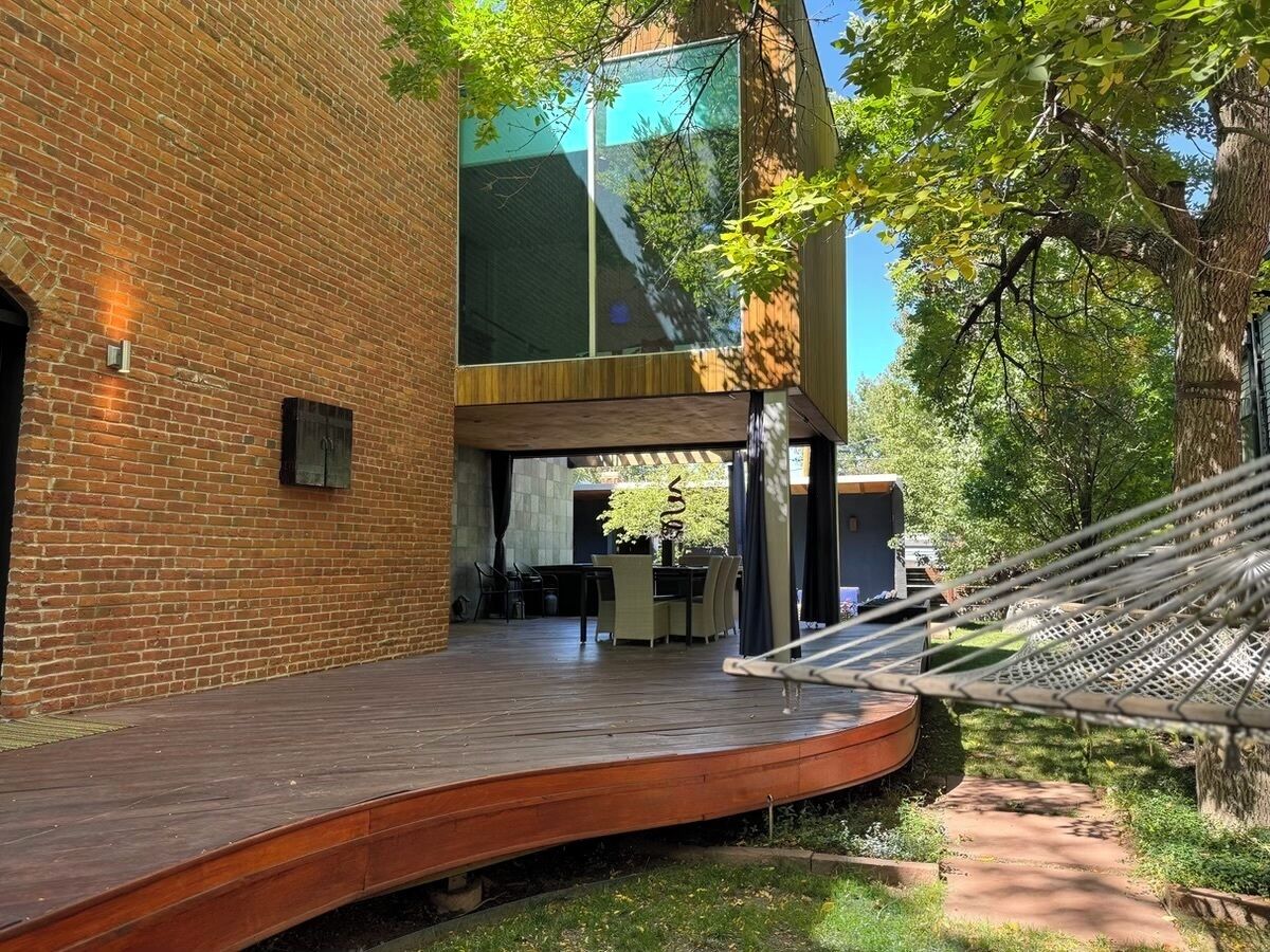 Un espace extérieur moderne doté d’une terrasse en bois, d’un hamac et d’un coin repas aménagé sous une structure en verre.
