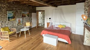 Un dormitorio rústico con vigas de madera a la vista, pared de piedra, cama con dosel, una mesita pequeña y una silla.