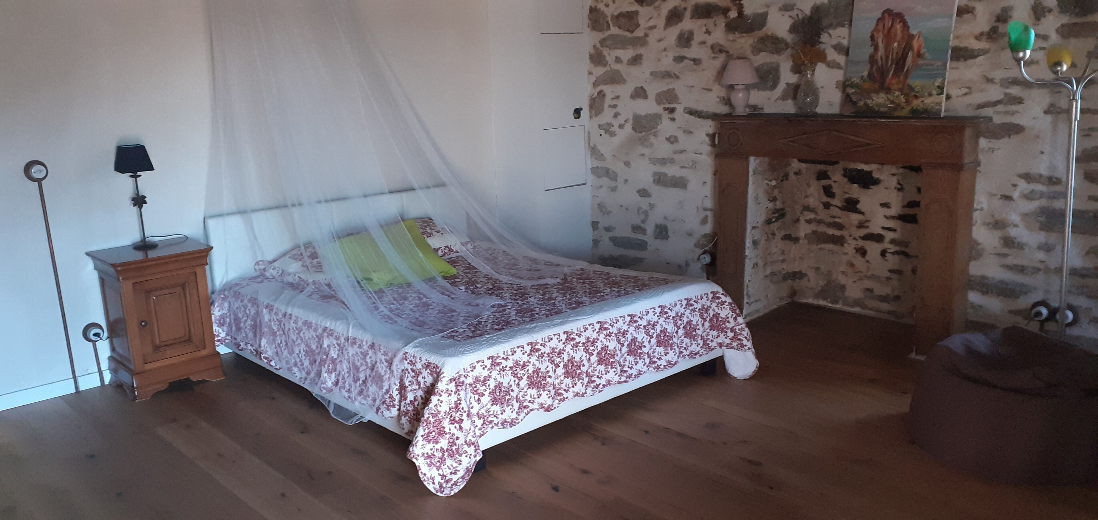 Une chambre à coucher avec un lit sous une moustiquaire, une table de chevet avec une lampe, une armoire en bois, un mur en pierre, une cheminée et une petite statue posée sur le manteau de la cheminée.