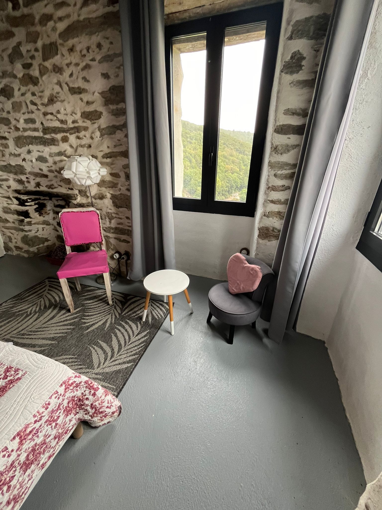 Une pièce avec un mur en pierre, une fenêtre donnant sur la verdure, une chaise rose, une petite table ronde et un lit recouvert d’une courtepointe à motifs.