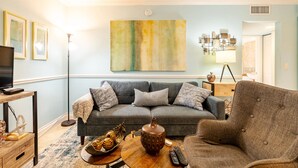 A living room with a grey sofa, a coffee table with a vase and decorative items, a television on a stand, and a large abstract painting on the wall. - Sunny Retreat: Cozy APT in WPB #2 (West Palm Beach)