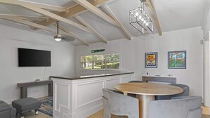 A modern kitchen with a central island, a dining area with a round table and chairs, a flat-screen TV mounted on the wall, and a window with blinds.