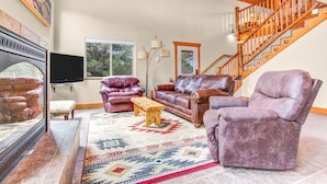 A spacious living room with a large TV, leather sofas, a wooden coffee table, and a patterned rug. A wooden staircase is visible in the background.