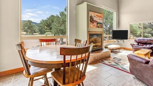A spacious room with a round wooden dining table and chairs, a fireplace, a television mounted on the wall, and a large window offering a view of the outdoors.