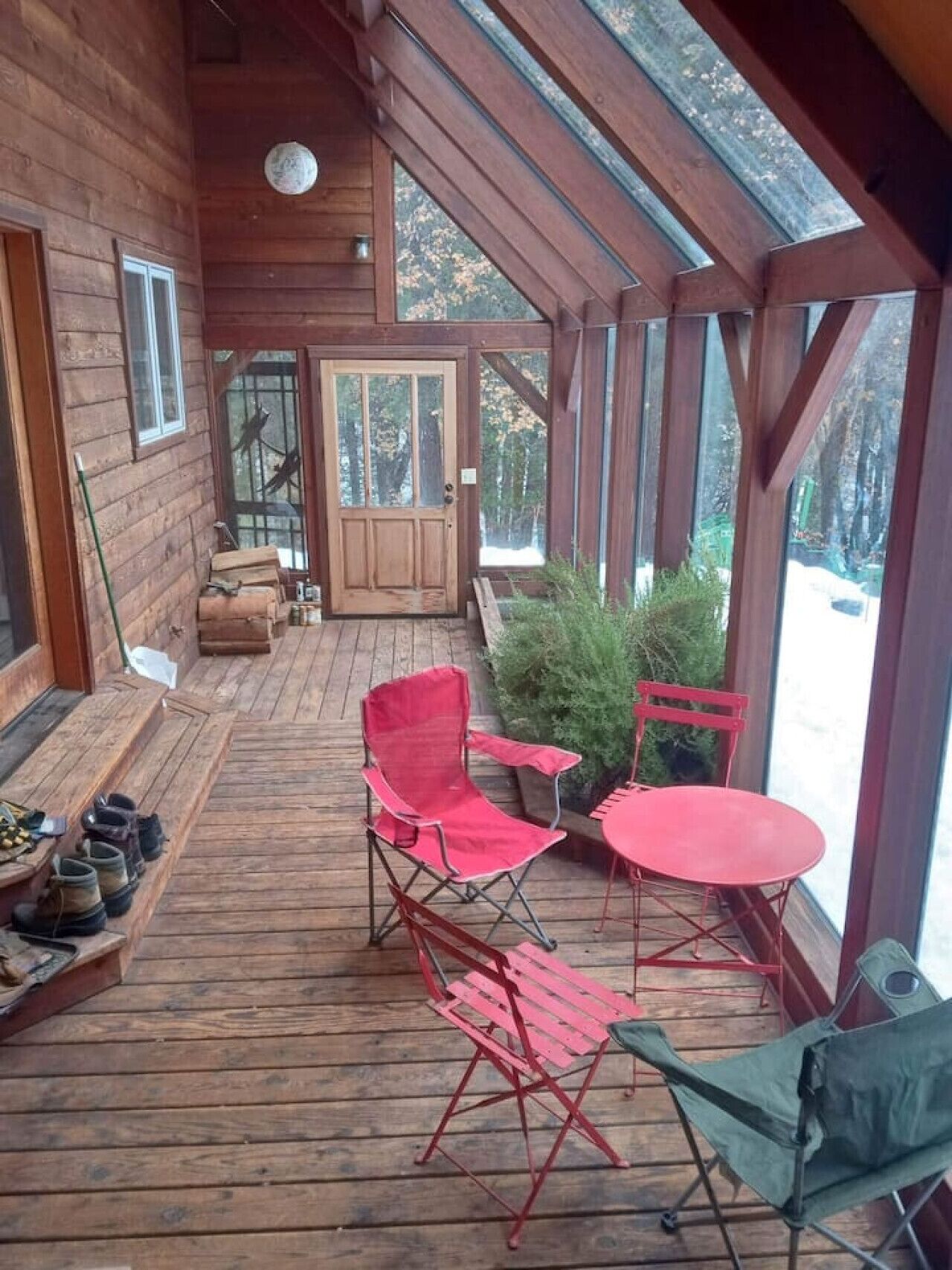 A wooden deck with red folding chairs and a table, a door with a glass panel, and a view of trees through the windows.