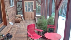 Une terrasse en bois avec des chaises pliantes rouges et une table, une porte avec un panneau de verre, et une vue sur les arbres à travers les fenêtres.