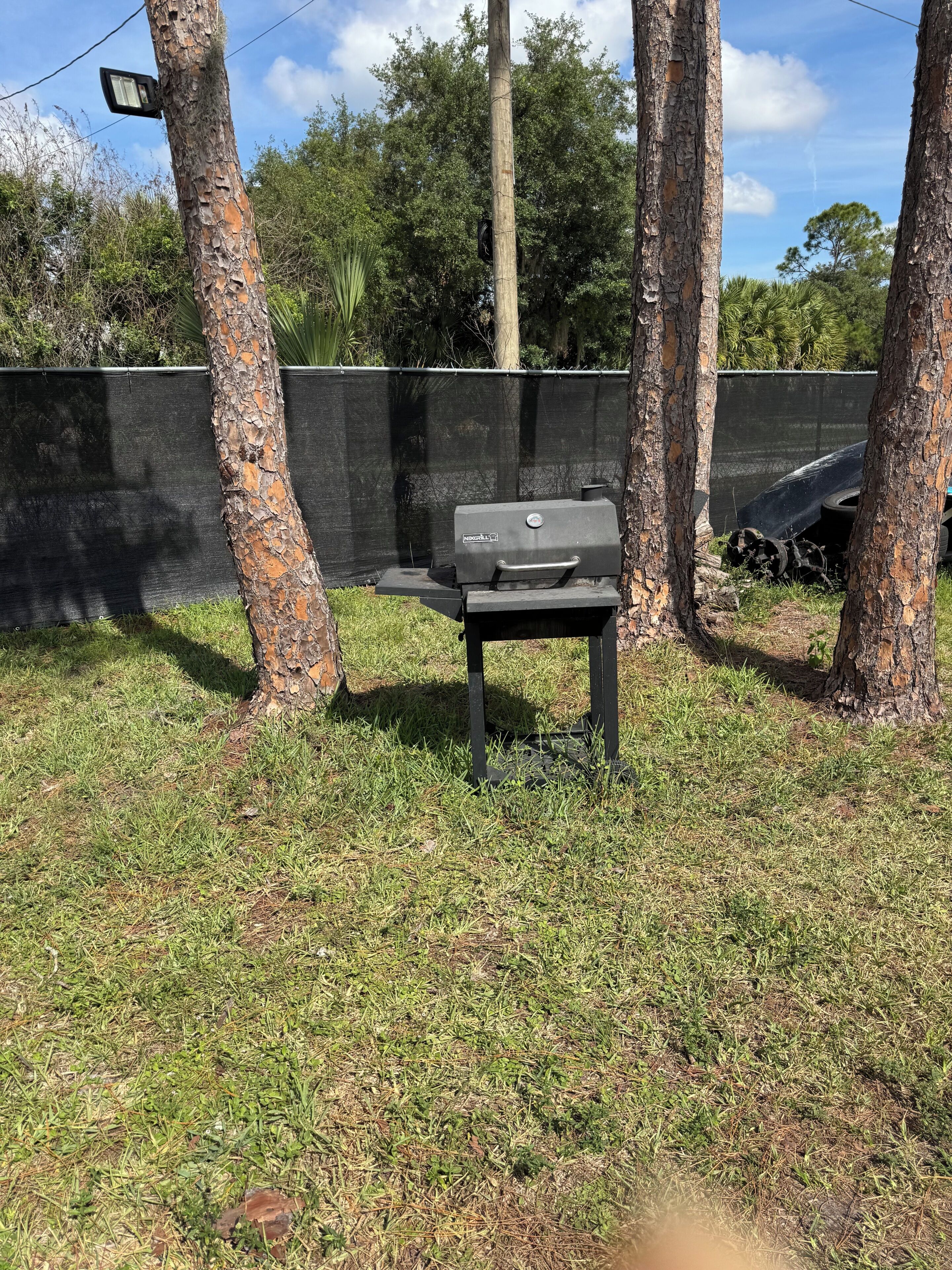 Una griglia per barbecue in una zona all'aperto erbosa, con alberi e una recinzione sullo sfondo.