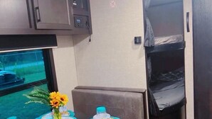 A compact RV interior with a kitchenette, a dining area with blue tableware, and a window with a view of greenery. - “RV Country Living” Glamping (Clewiston)