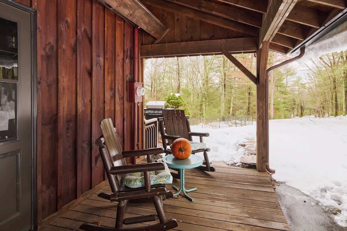 A wooden porch with two rocking chairs and a small table, a grill, and a view of a snowy forest.