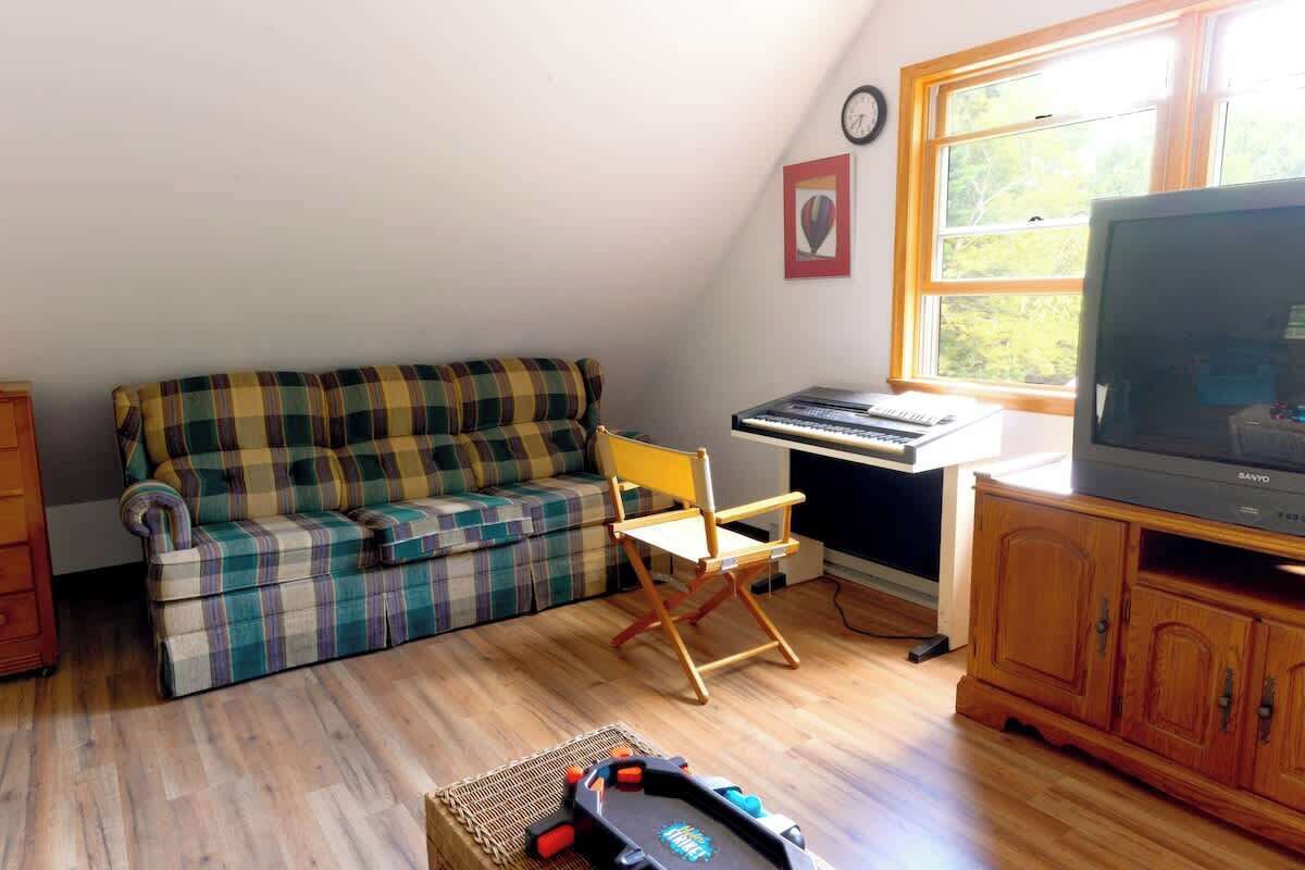 Ein Zimmer mit einem Karo-Sofa, einem Holztisch mit Tastatur, einem Fernseher und einem Fenster mit Blick auf Bäume.