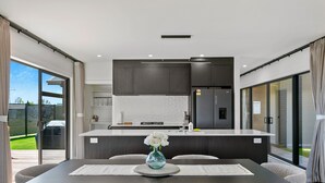 A modern kitchen with dark cabinetry, a central island, and a dining area with a table and chairs. - Stylish New Retreat in Taupo (Taupo)