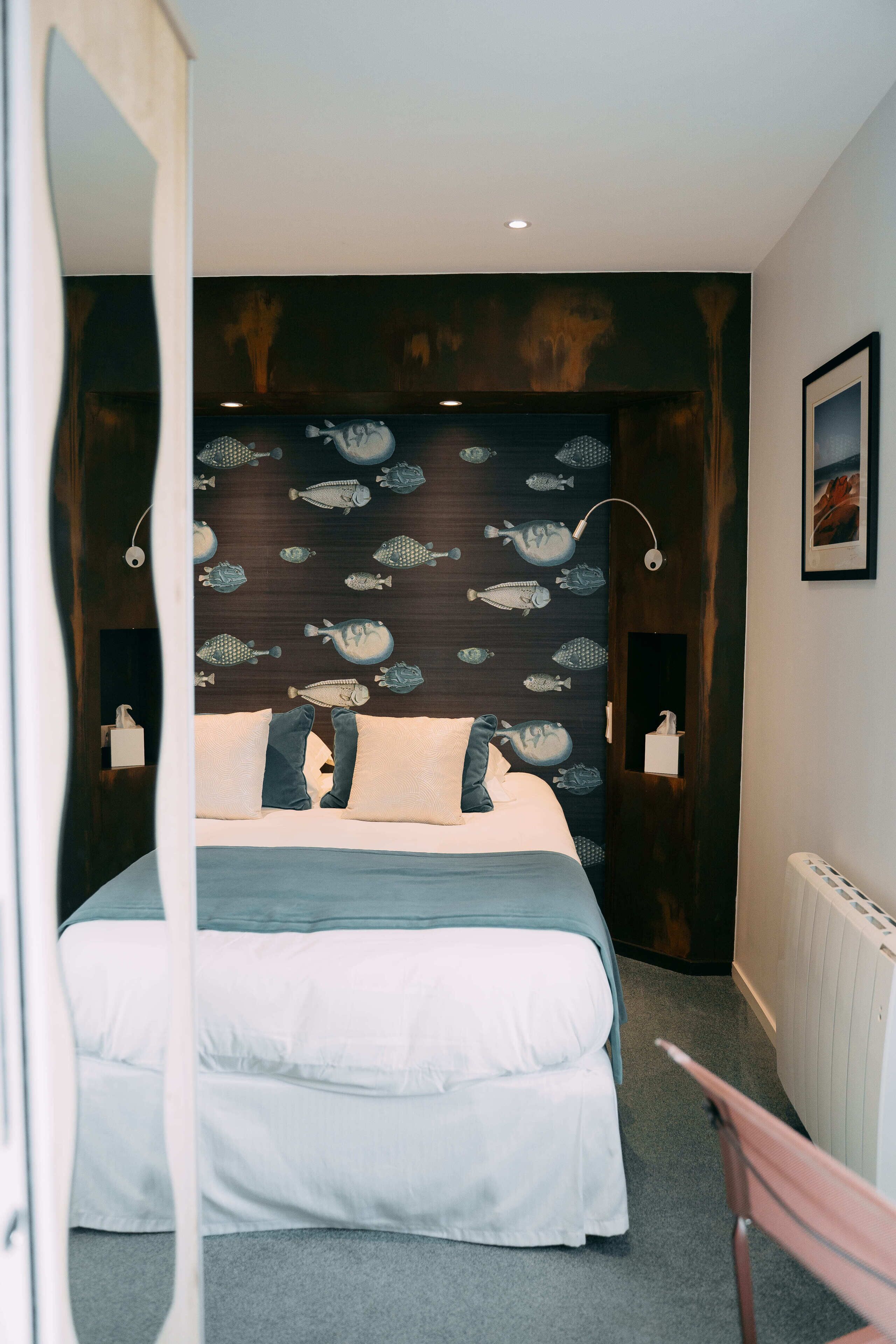 Une chambre à coucher avec un lit, des oreillers, une tête de lit ornée de motifs de poissons, des lampes de chevet et un tableau encadré accroché au mur.