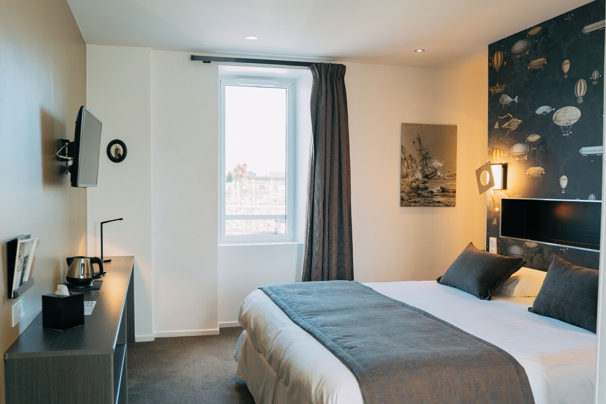 Une chambre d’hôtel moderne dotée d’un grand lit, d’une télévision à écran plat, d’un bureau avec une bouilloire et d’une fenêtre avec des rideaux.