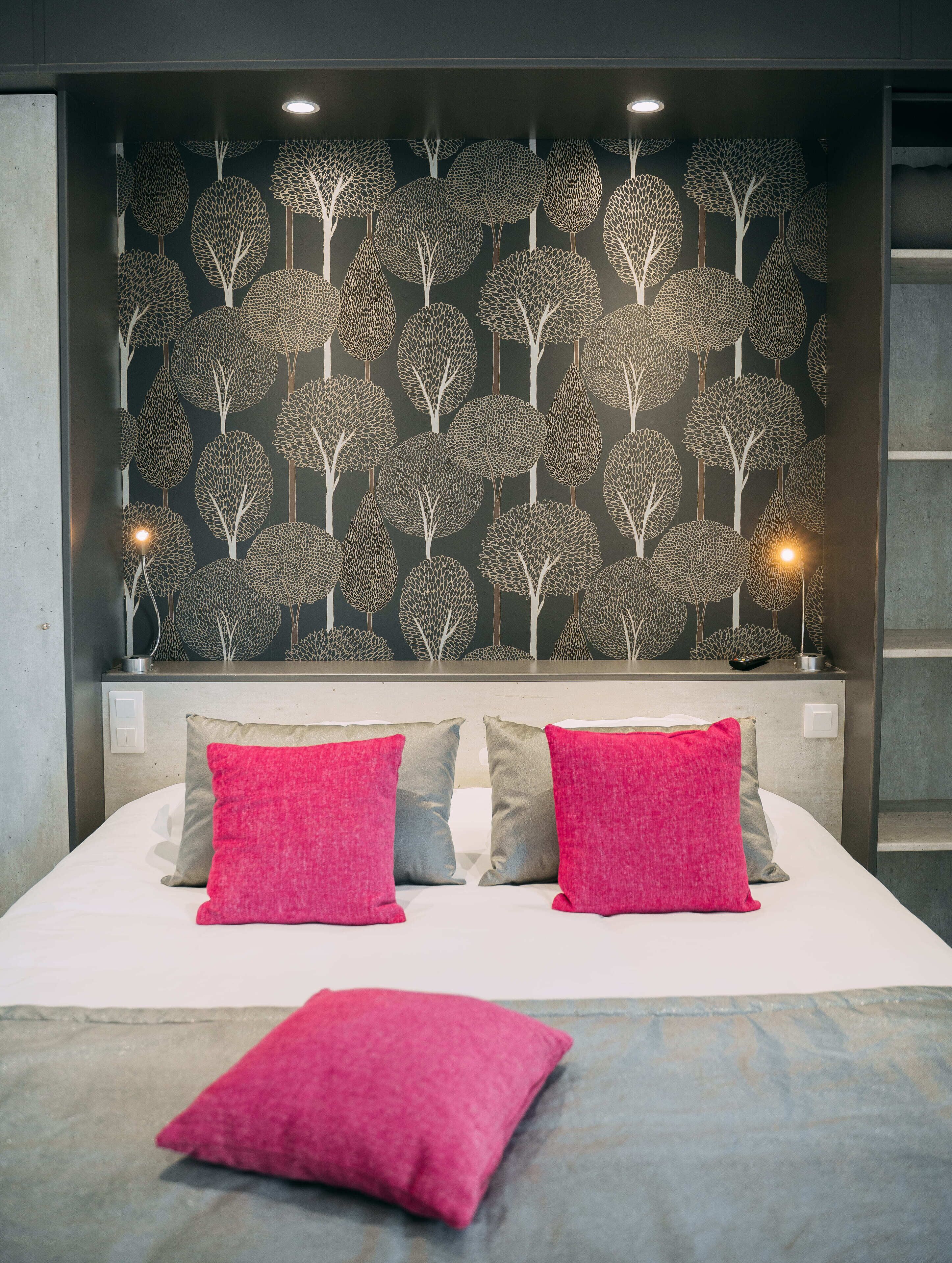 Una cama con dos almohadas rosas y una almohada gris, un cabecero gris y una pared con un diseño de árbol.