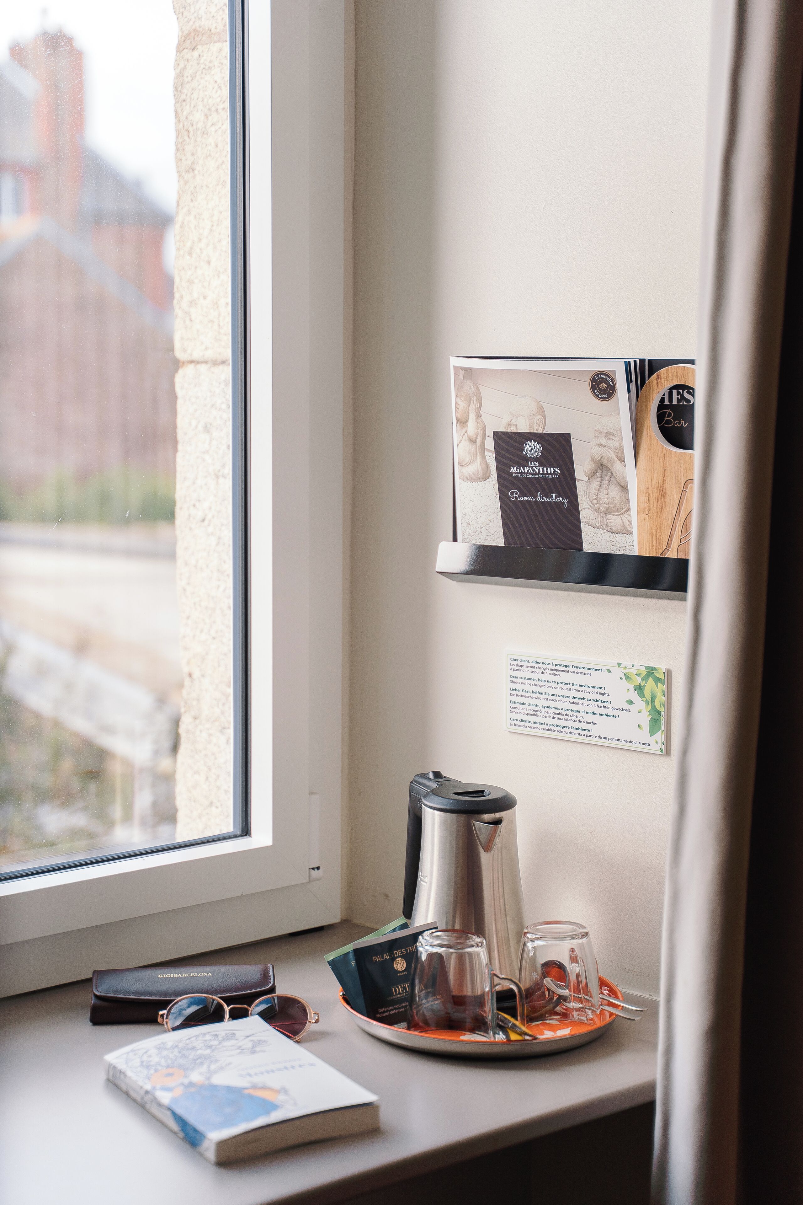 A window view of a cityscape, a coffee machine, a tray with glasses and a book, and a shelf with a menu and a sign.