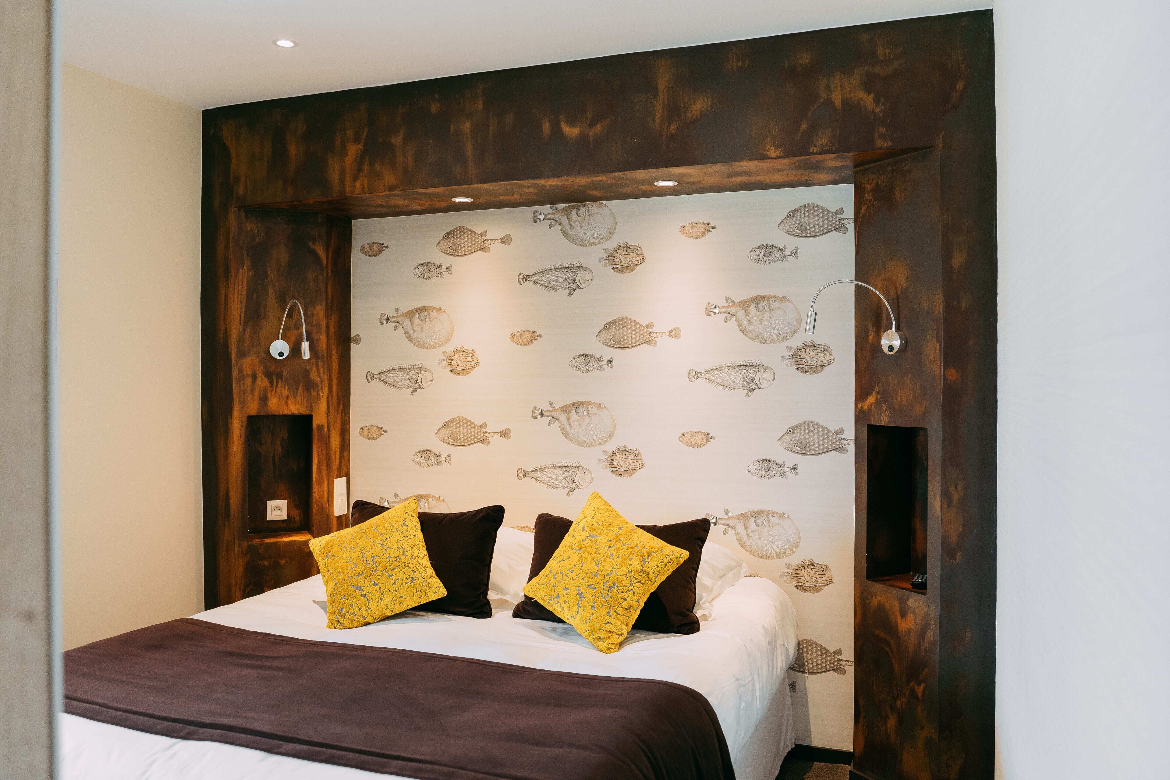 A bedroom with a bed, two yellow pillows, a brown bedspread, and a wall with fish patterns.