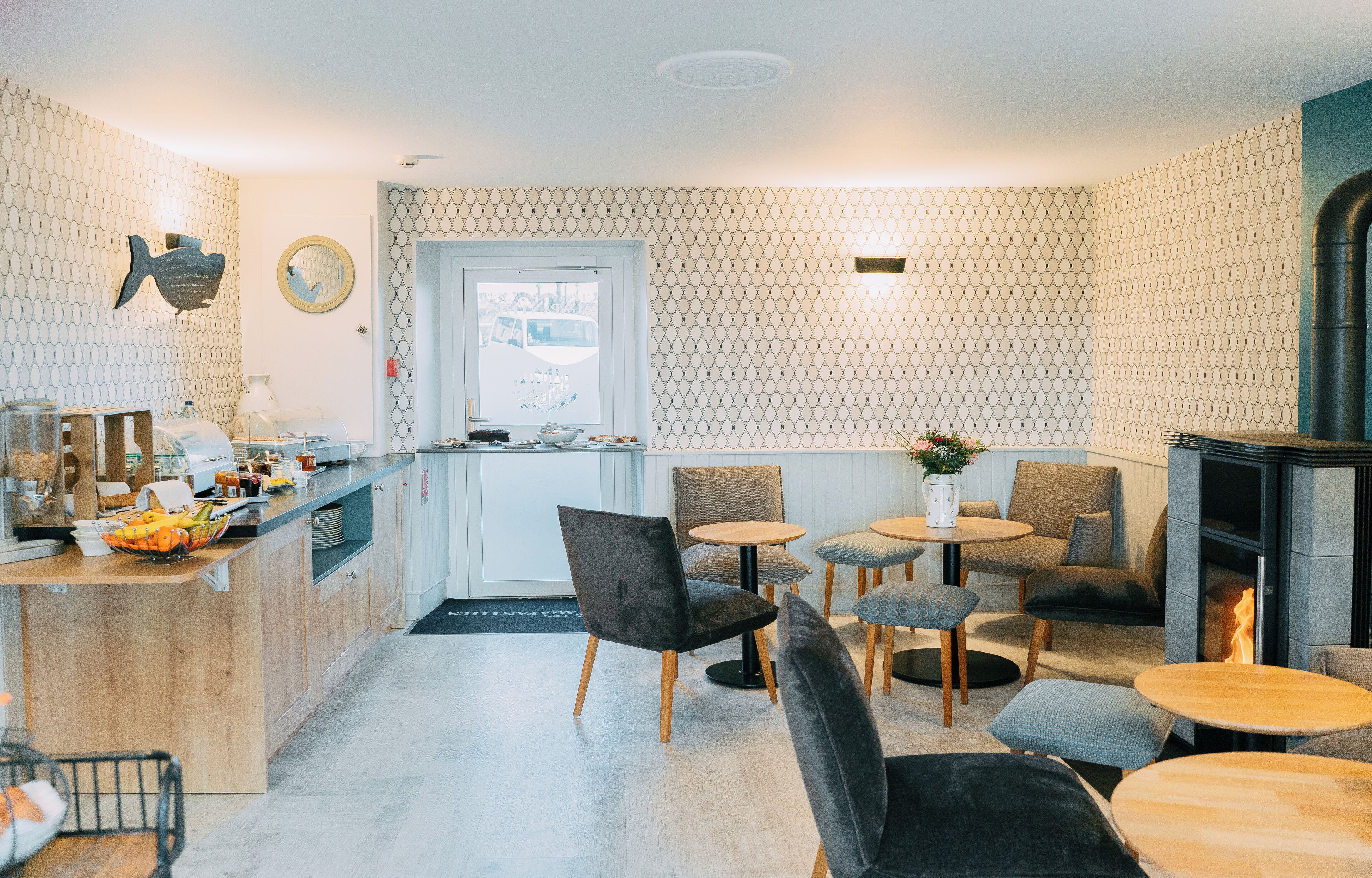 Une cuisine moderne avec un plan de travail en bois, un évier et une fenêtre. Des chaises et des tables sont disposées autour d’un espace central où se trouve un vase de fleurs.
