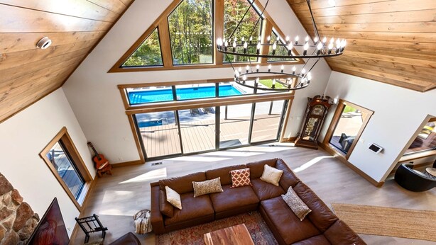 A spacious living area with a large brown leather sofa, a wooden coffee table, and a view of a swimming pool through large windows. - Lazaj Luxury Lodge — Heated Pool,Hot Tub, Game Room,Sleeps 16 Near Hocking Hills (Logan)