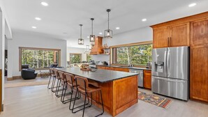 A modern kitchen with a large island, wooden cabinets, stainless steel appliances, and large windows offering a view of trees. - Lazaj Luxury Lodge — Heated Pool,Hot Tub, Game Room,Sleeps 16 Near Hocking Hills (Logan)