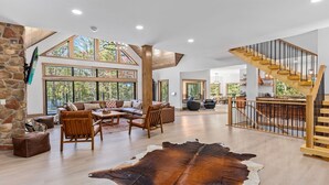 A spacious living area with a stone wall, large windows, a wooden staircase, and a leather rug. - Lazaj Luxury Lodge — Heated Pool,Hot Tub, Game Room,Sleeps 16 Near Hocking Hills (Logan)