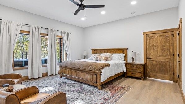 Un dormitorio con cama de madera, ventilador de techo, mesita de noche, armario de madera y una ventana grande con cortinas.