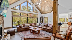 A spacious living room with a large L-shaped sectional sofa, a wooden coffee table, and a large window offering a view of trees. - Lazaj Luxury Lodge — Heated Pool,Hot Tub, Game Room,Sleeps 16 Near Hocking Hills (Logan)