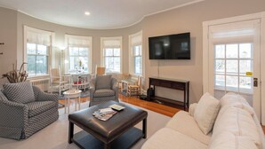 A living room with a white sofa, patterned armchairs, a coffee table, a TV mounted on the wall, and large windows with white curtains. - Gorgeous Spacious Victorian Cape Cod Beach View Retreat (Falmouth)