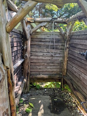 Une structure en bois, dotée d’une pomme de douche en métal et d’un tuyau, entourée d’une clôture en bois et de verdure.