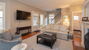 A living room with a sofa, a coffee table, and a TV mounted on the wall. - Gorgeous Spacious Victorian Cape Cod Beach View Retreat (Falmouth)
