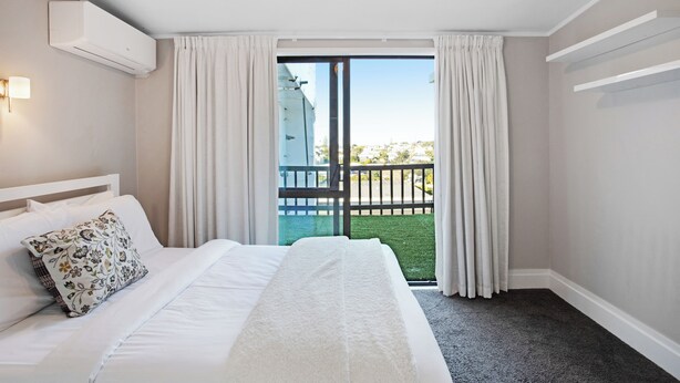 Quarto de hotel com cama, cortinas brancas, ar condicionado e vista para uma varanda com grade.